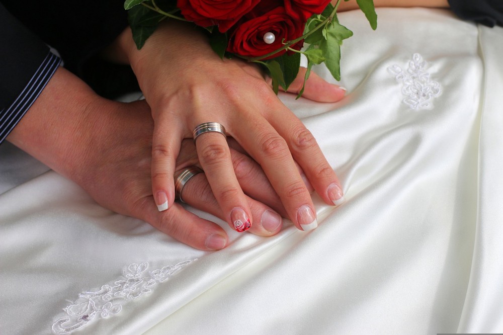 The hands of the bride and the groom one another on the bride's dress, while both wedding bands wearing