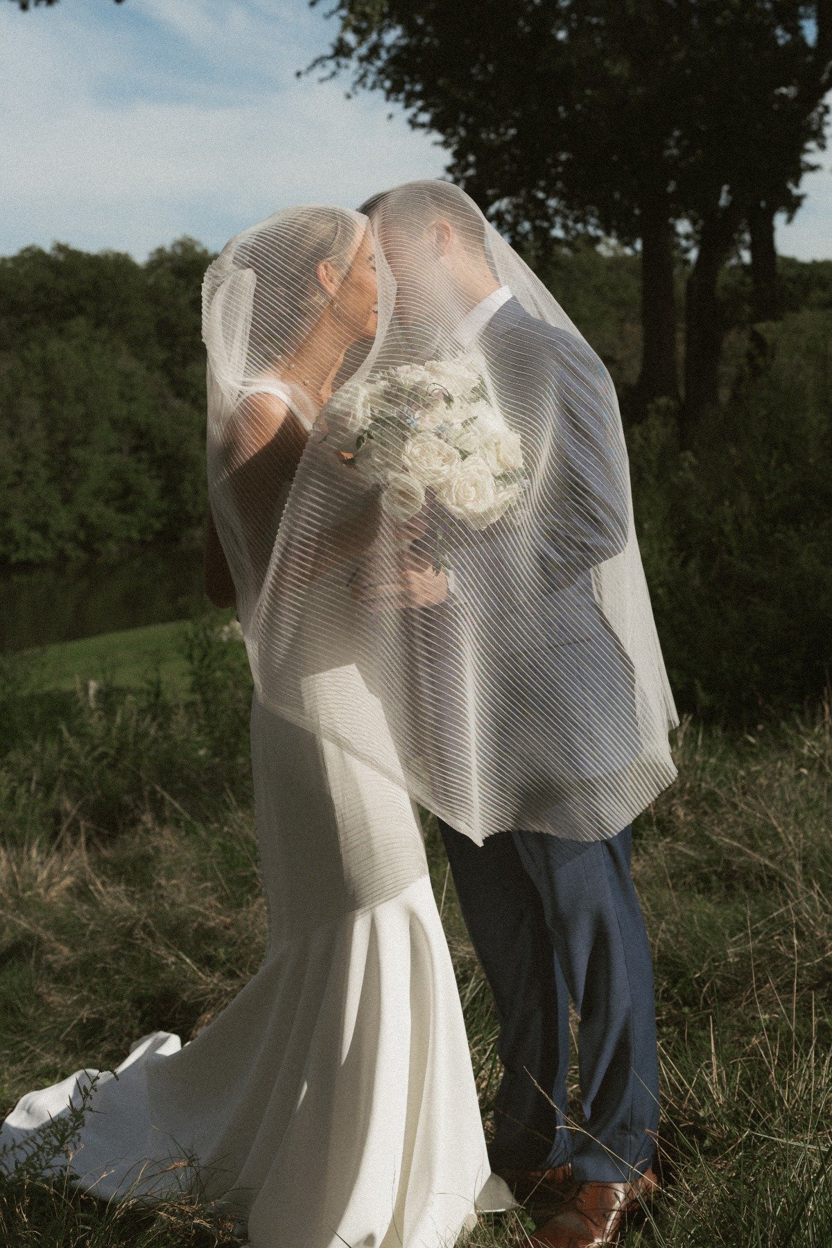 bride and groom kiss in field under a pleated veil